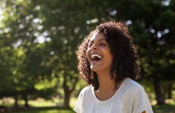 Woman with curly hair laughing outdoors, surrounded by trees.